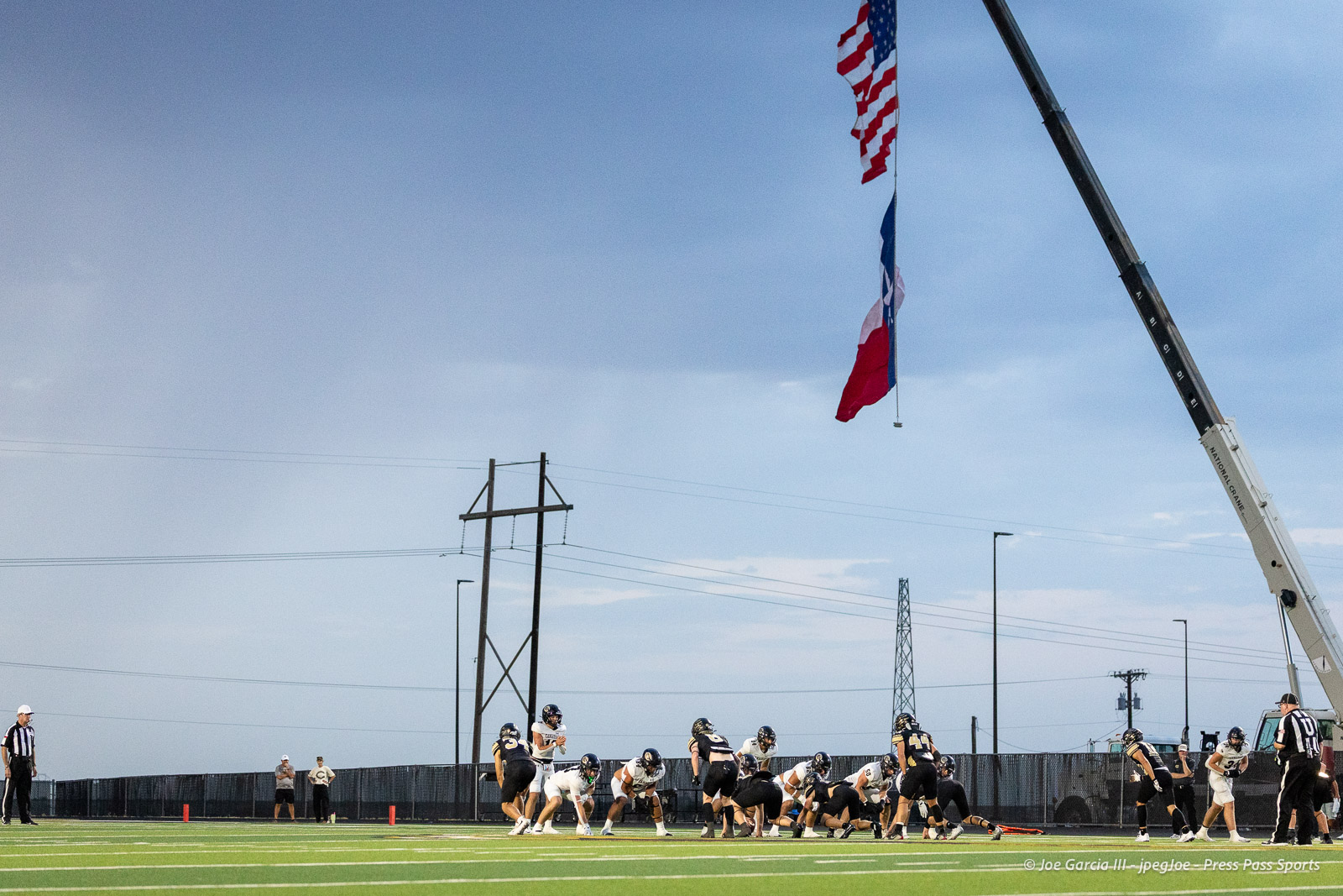 Football players line up at the line of scrimmage on a sunny field with an American flag and Texas flag raised high on a crane overhead.