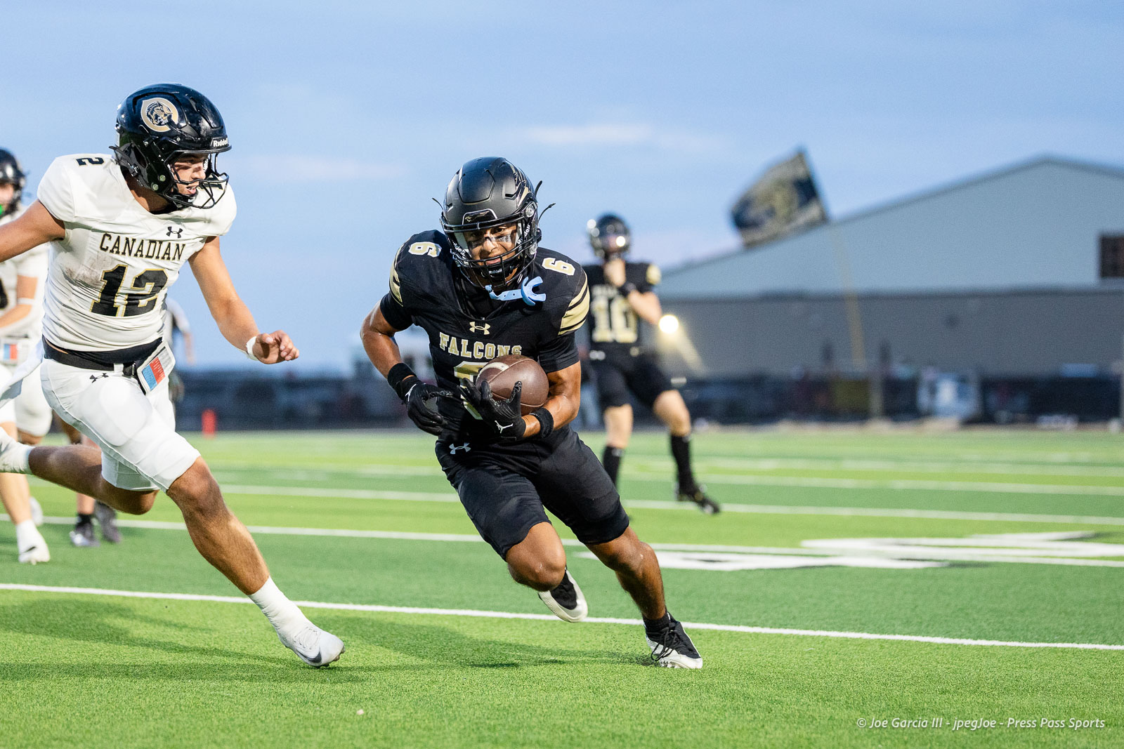 Falcons player in a black jersey runs with the ball, pursued by a Canada team player in white on a green turf field.