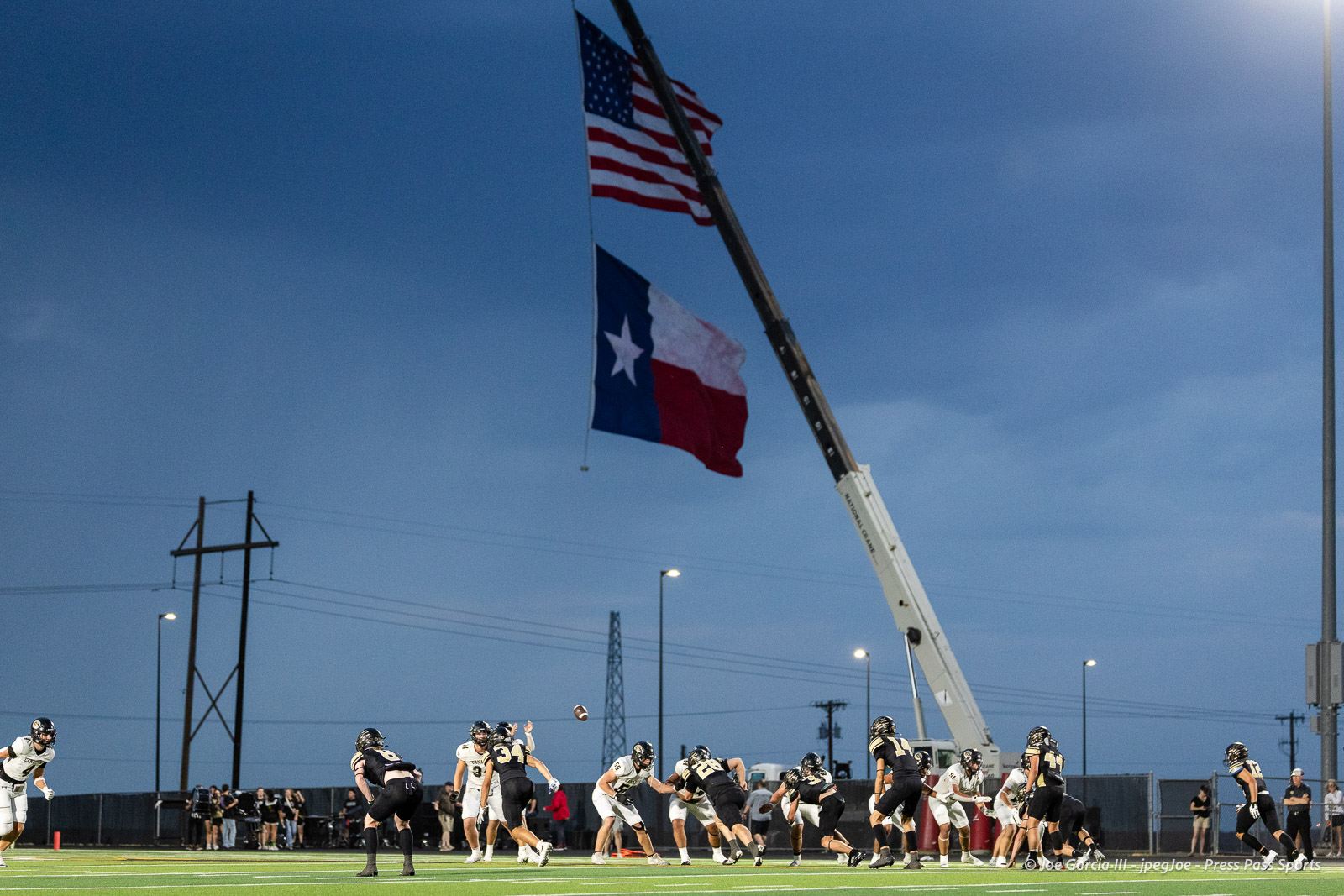 Football game on a lit field as a tall crane hoists large American and Texas flags above the players.