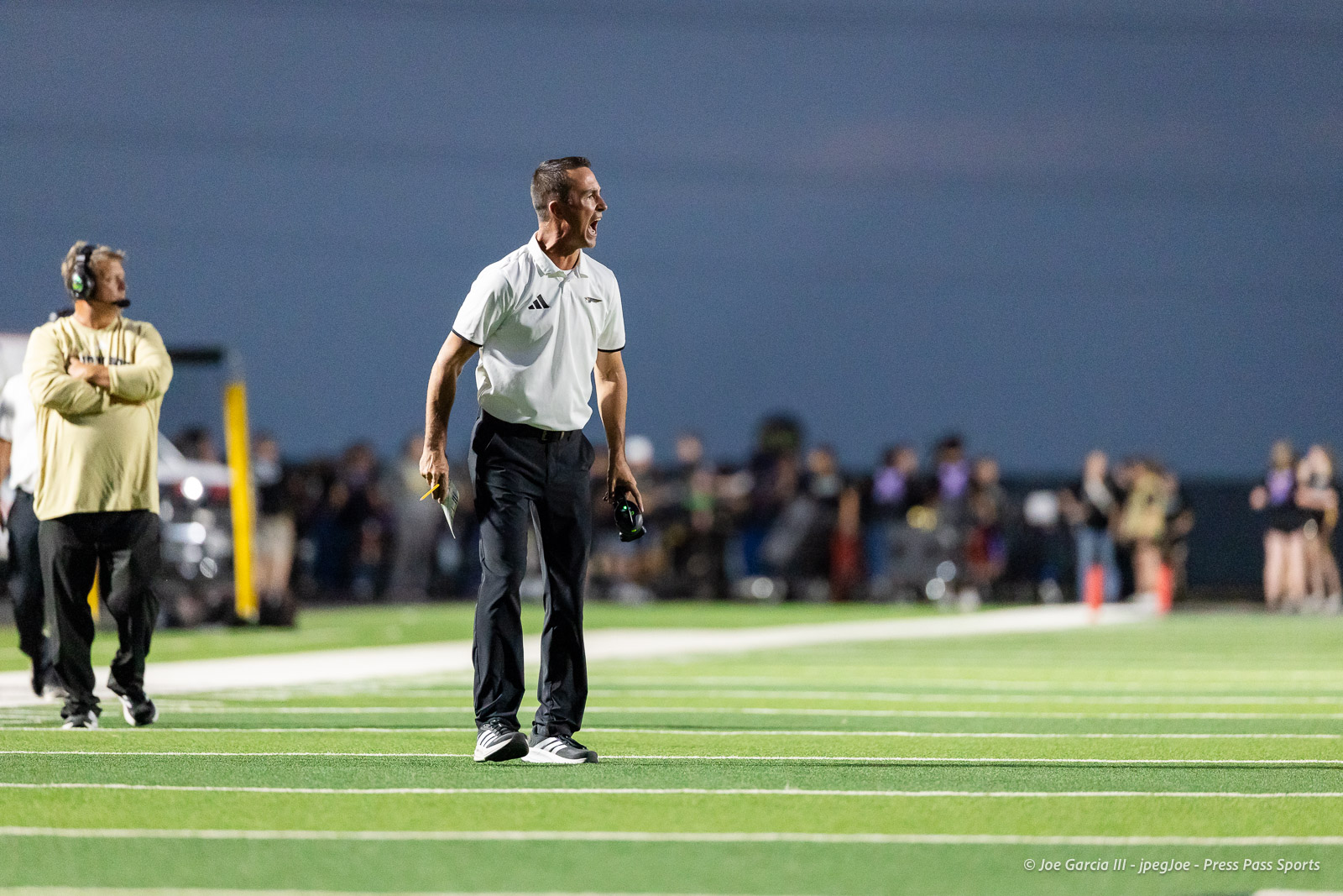 Coach on a football field shouting instructions, holding a whistle and a clipboard, with blurred spectators in the background at dusk.
