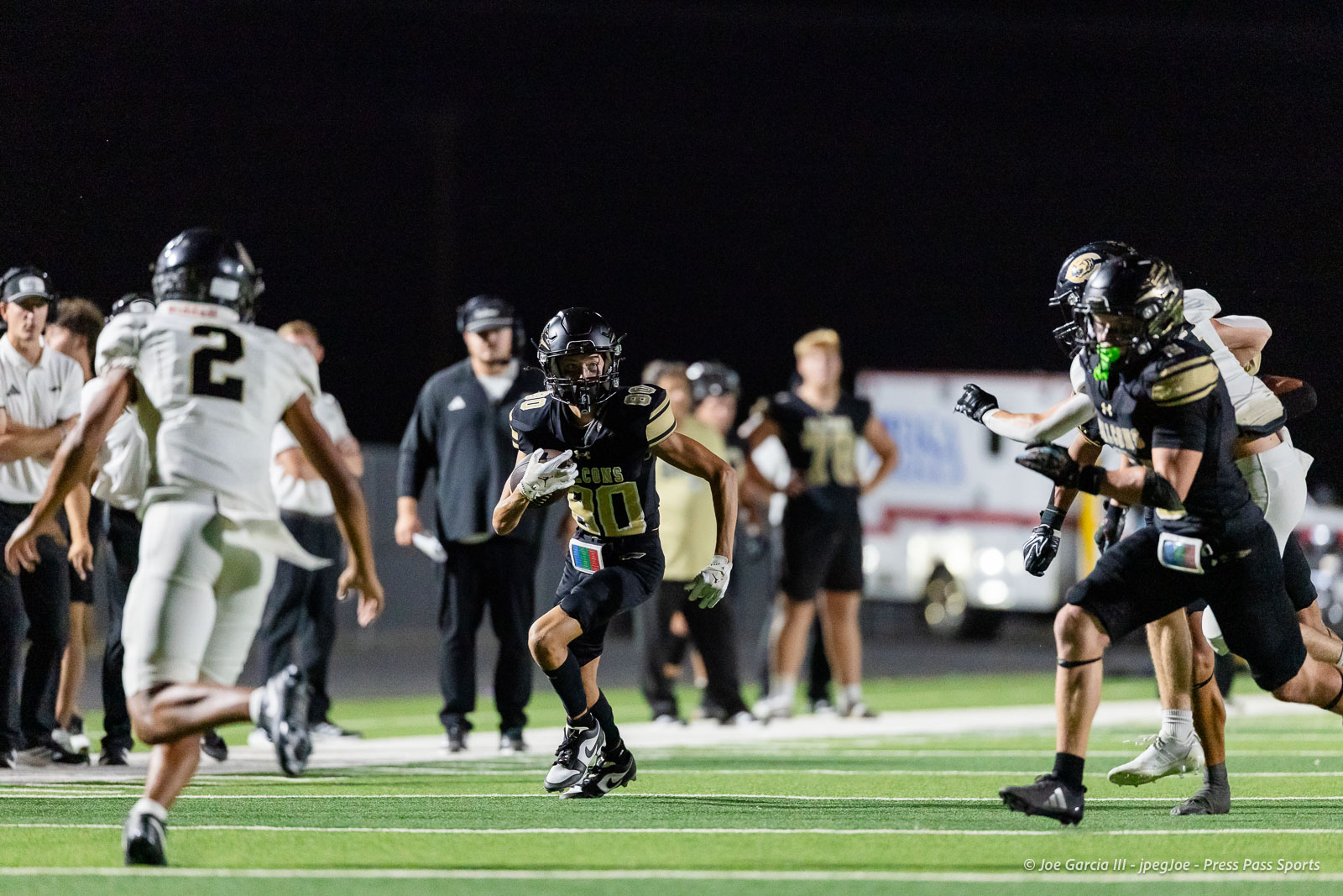 Football game at night: a player in black #80 carries the ball while defenders approach on the green turf under lights.