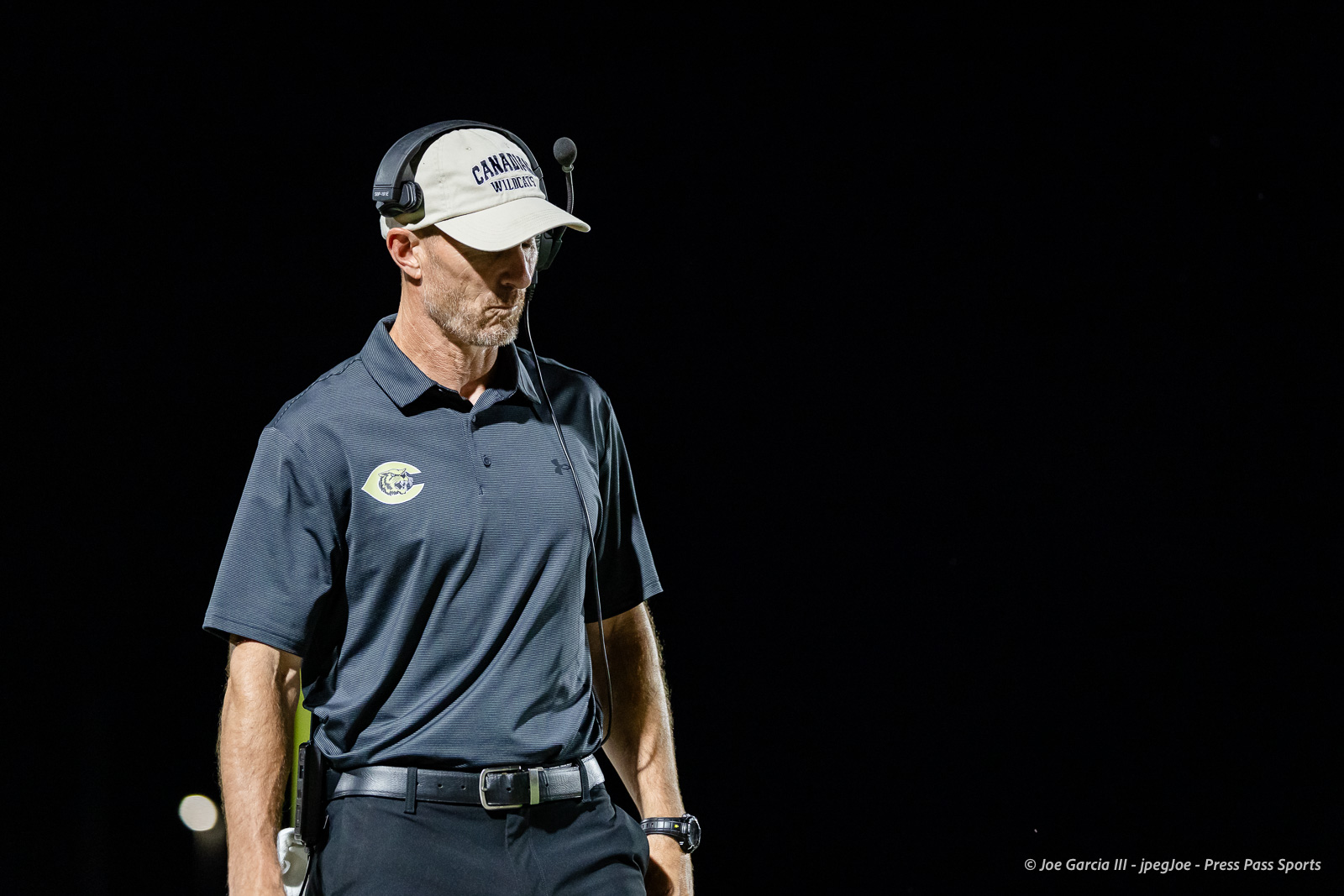 Male coach wearing a headset and white cap, standing against a dark night background in a gray polo with a logo.