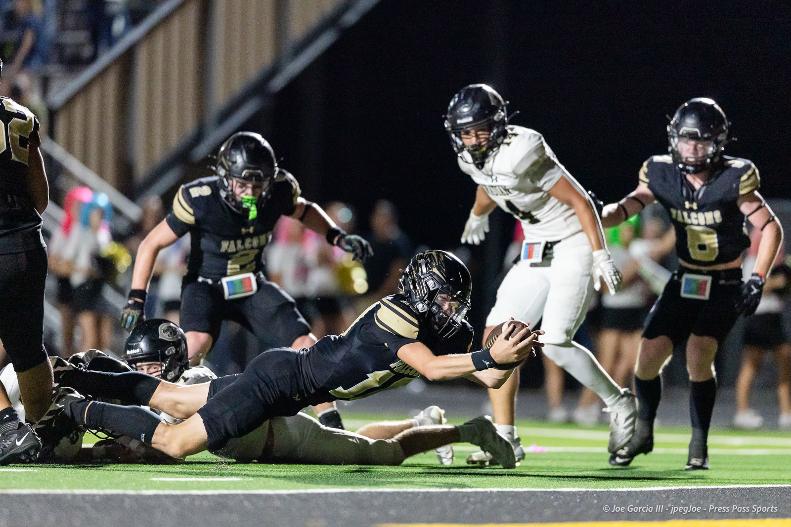 A football player in black and gold dives forward to catch a ball while others chase during a night game on a turf field.