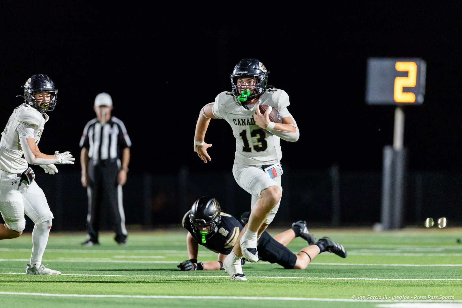 Football player wearing number 13 runs with the ball during a night game on a lit field; other players and a referee in the background.