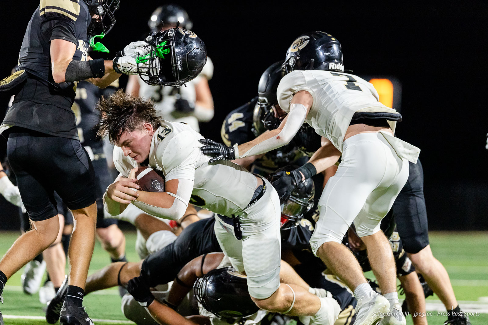 Football scene: a white-jersey running back grips the ball as black-uniform defenders converge on a nighttime turf play.