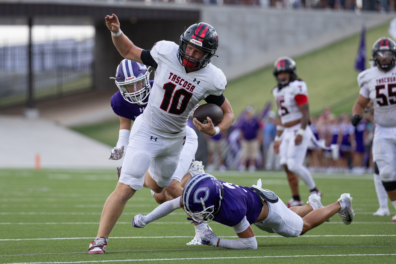 THS-CHS-FB-14 | Press Pass Sports White-jersey football player, number 10, runs with the ball as a purple-clad defender dives to tackle him on a green field.