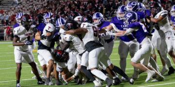 Football players collide at the line of scrimmage during a nighttime game, white and purple uniforms in a tight scrum.