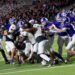 Football players collide at the line of scrimmage during a nighttime game, white and purple uniforms in a tight scrum.
