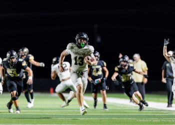 canadian-vs-bushland-football-jpegJoe-pps-23 | Press Pass Sports White-jersey football player (#13) for Canadian hops forward with ball at night, pursued by black-jersey opponents on a bright turf field, crowd blurred in background.