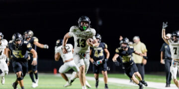 White-jersey football player (#13) for Canadian hops forward with ball at night, pursued by black-jersey opponents on a bright turf field, crowd blurred in background.