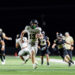 canadian-vs-bushland-football-jpegJoe-pps-23 | Press Pass Sports White-jersey football player (#13) for Canadian hops forward with ball at night, pursued by black-jersey opponents on a bright turf field, crowd blurred in background.