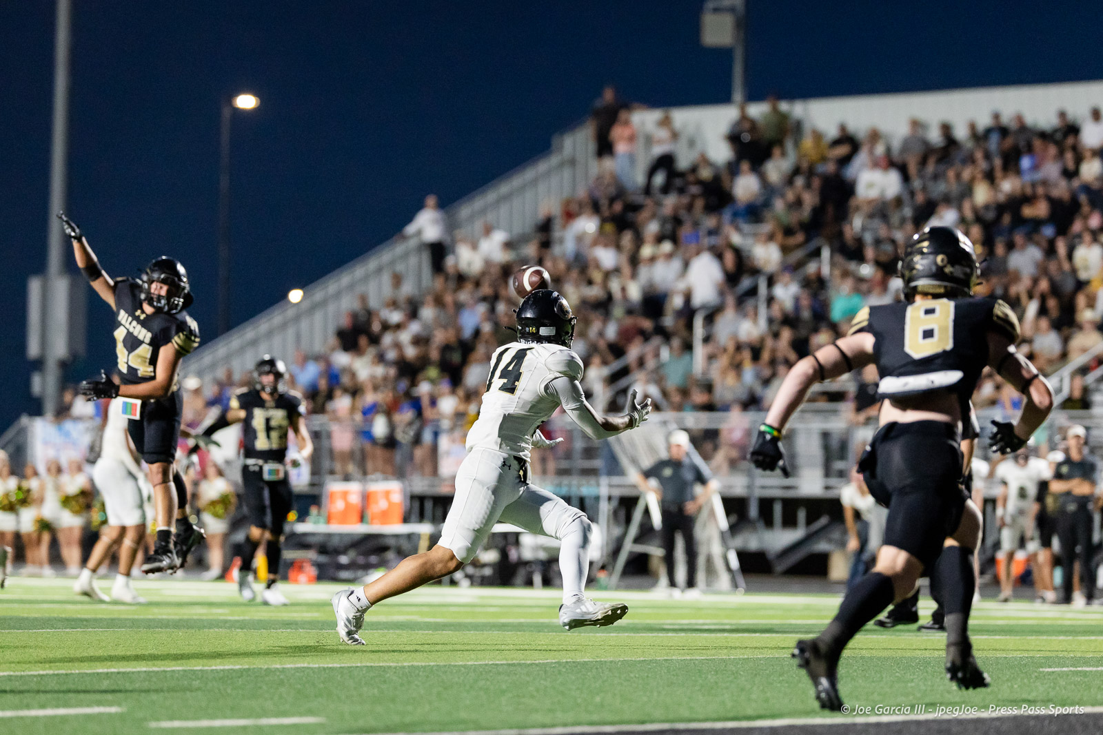 Action-packed夜 football scene: a white-jersied player (#14) runs toward the end zone as the football arcs in the air, chased by dark-uniform defenders with a full stands crowd in the background.