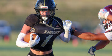 Female lacrosse player in a black jersey (#7) running with the ball, wearing a helmet and gloves during a game, defender approaching