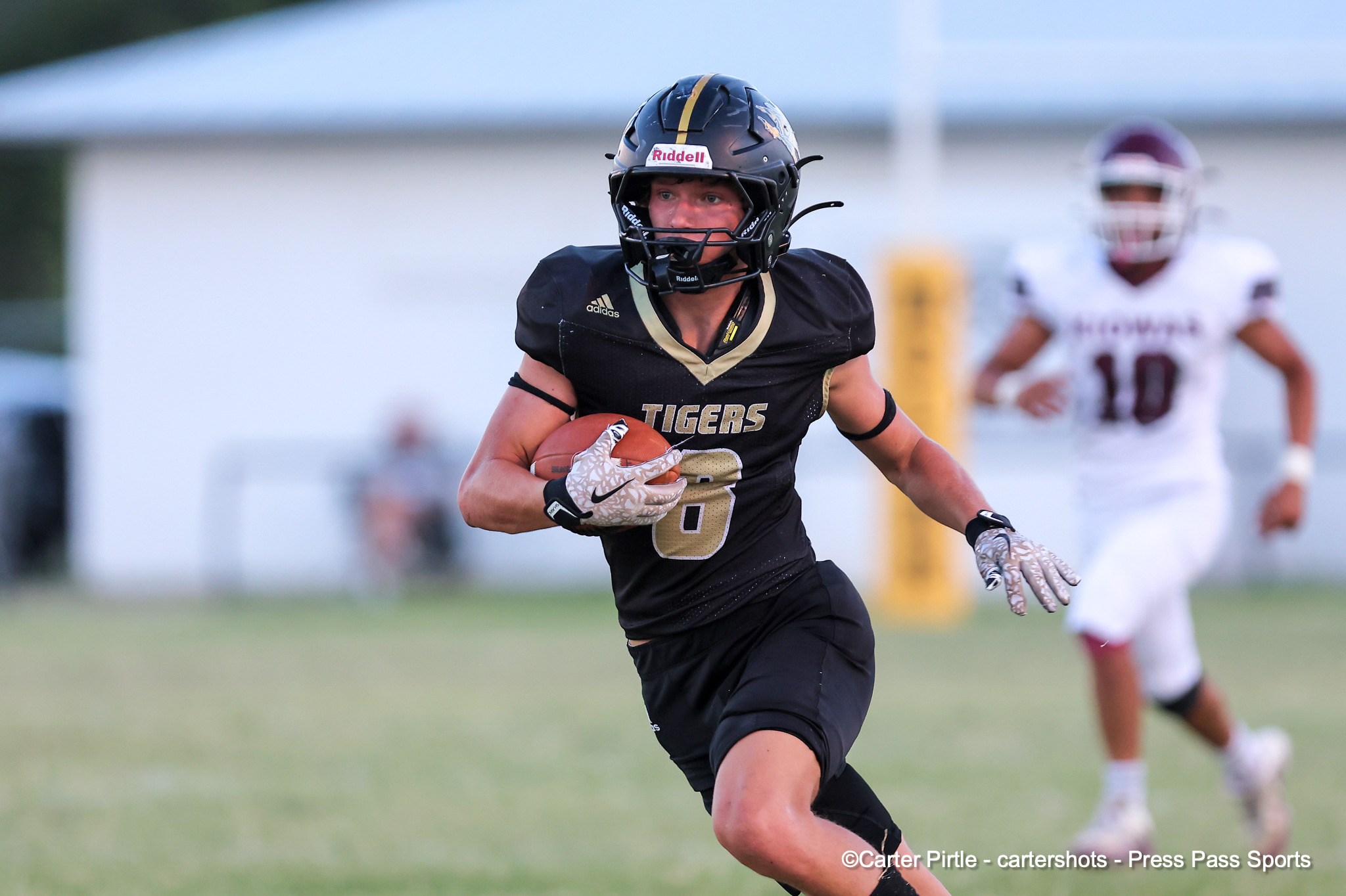Football player in black uniform carrying a ball, sprinting on the field during a game with a pursuing opponent in the background