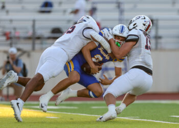 Football players in white tackle a blue-and-gold opponent during a game on a green field with stands in the background.
