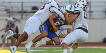 Football players in white tackle a blue-and-gold opponent during a game on a green field with stands in the background.
