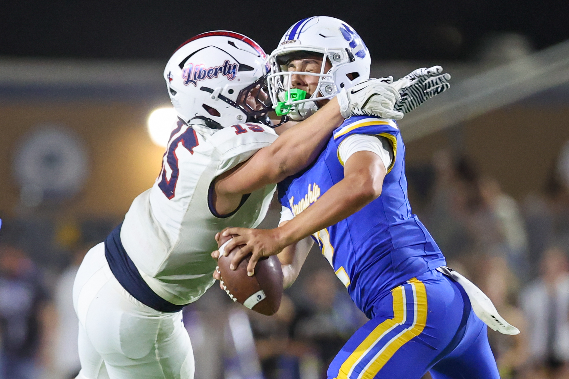Seabolt Chase-18033 | Press Pass Sports Two football players collide in midair during a play, one in a white uniform with a red Liberty helmet and the other in a blue and yellow uniform, both reaching for the ball.