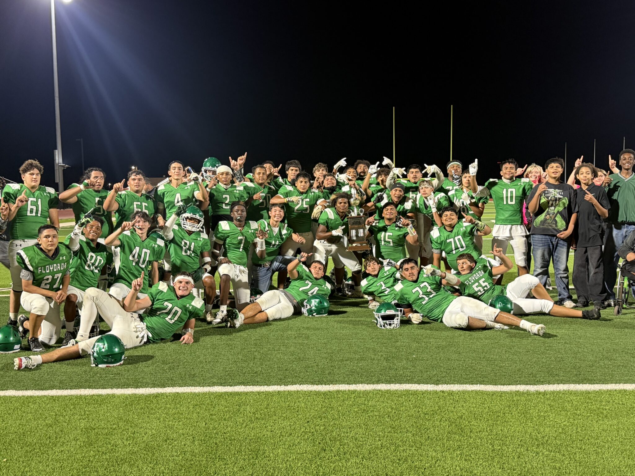 IMG_6943 | Press Pass Sports Group of football players in green uniforms posing on a field at night, with goalposts in the background and stadium lights overhead.