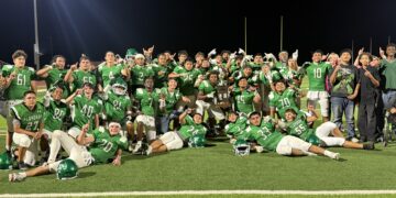Group of football players in green uniforms posing on a field at night, with goalposts in the background and stadium lights overhead.