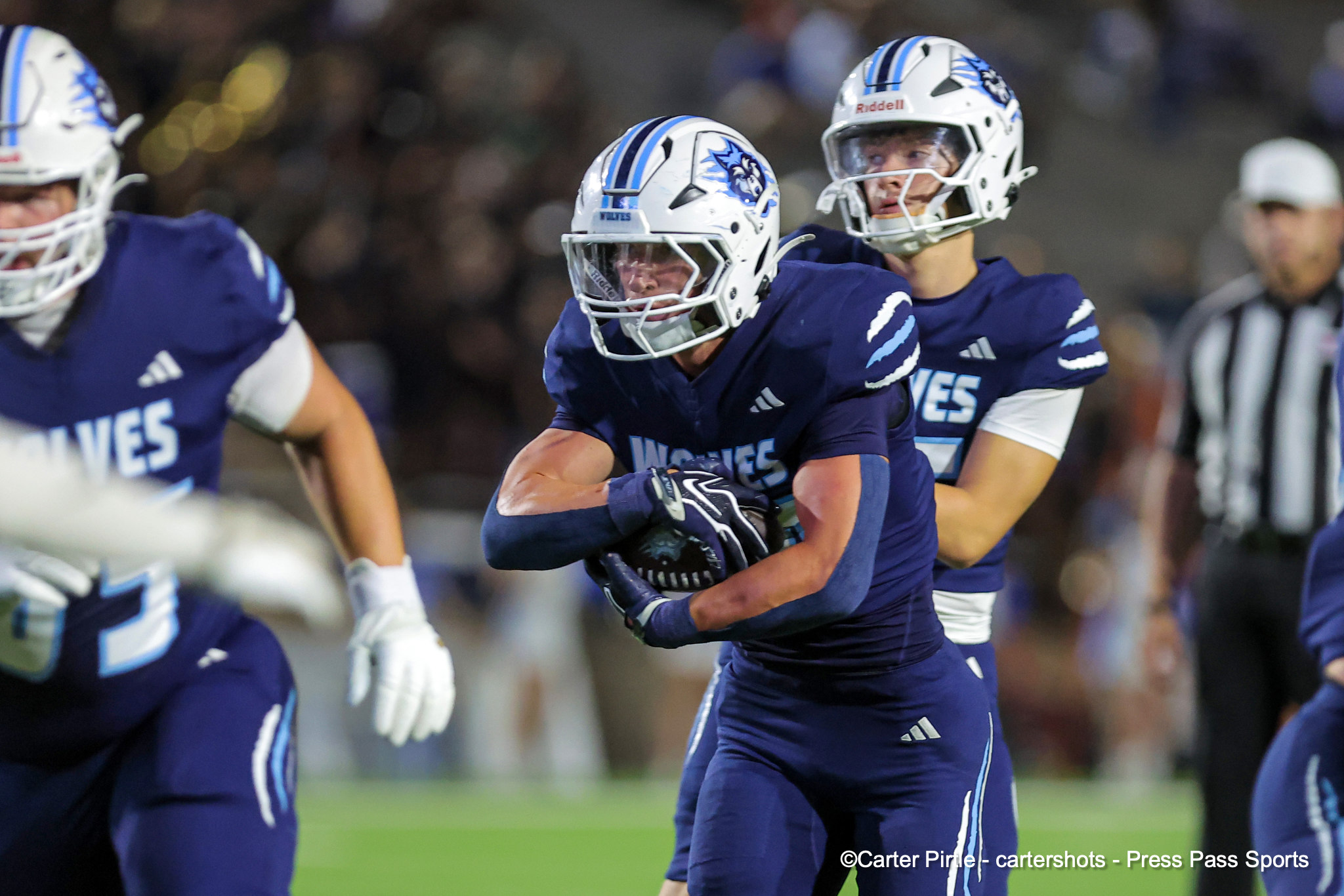 Football players in blue uniforms on a green field; central player in a white helmet carries the ball as teammates close in behind him.