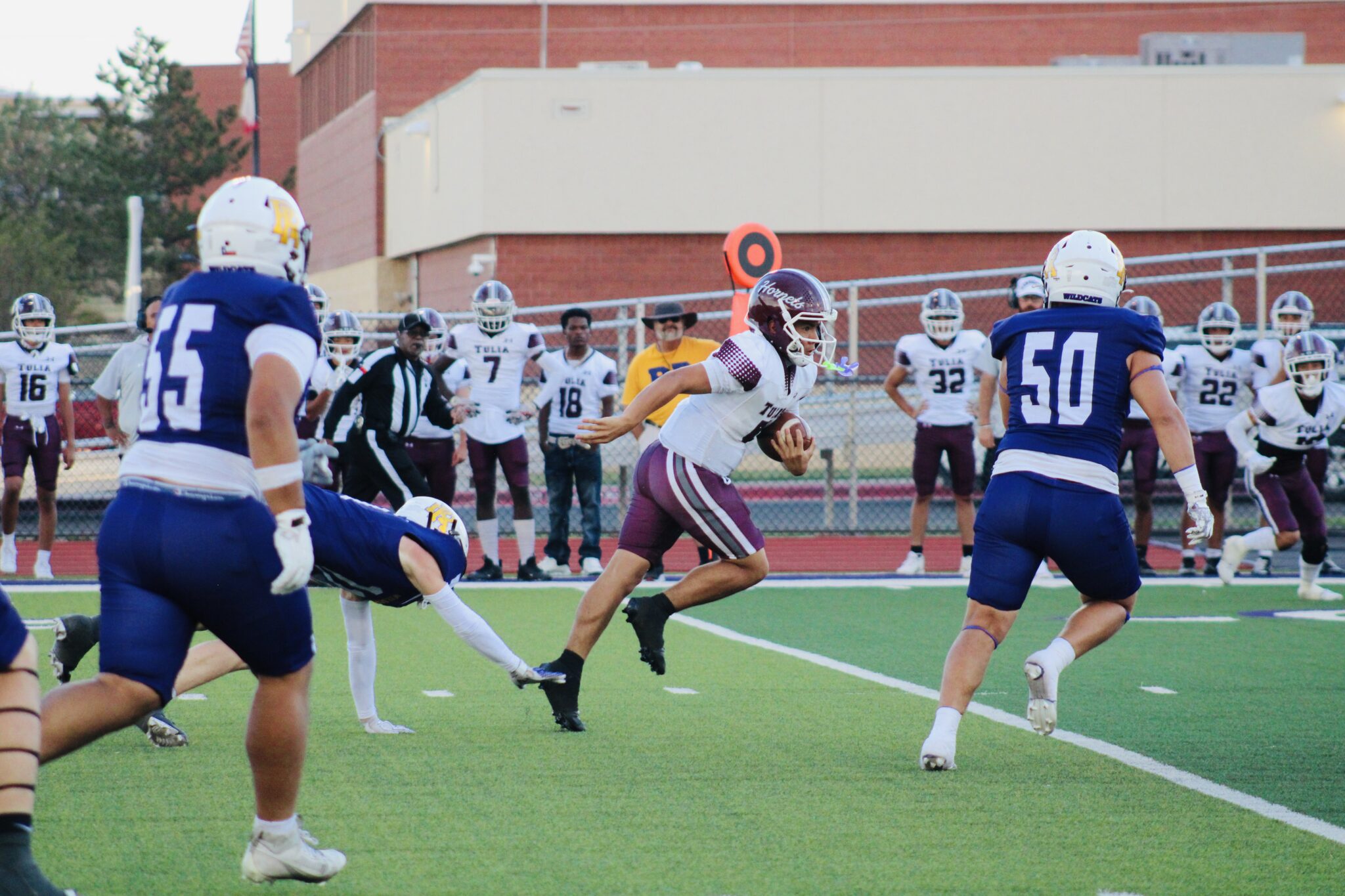 IMG_2528 | Press Pass Sports Football game action: a ball carrier in maroon and white sprints past blue-jersey defenders on a bright turf field by a chain-link fence.