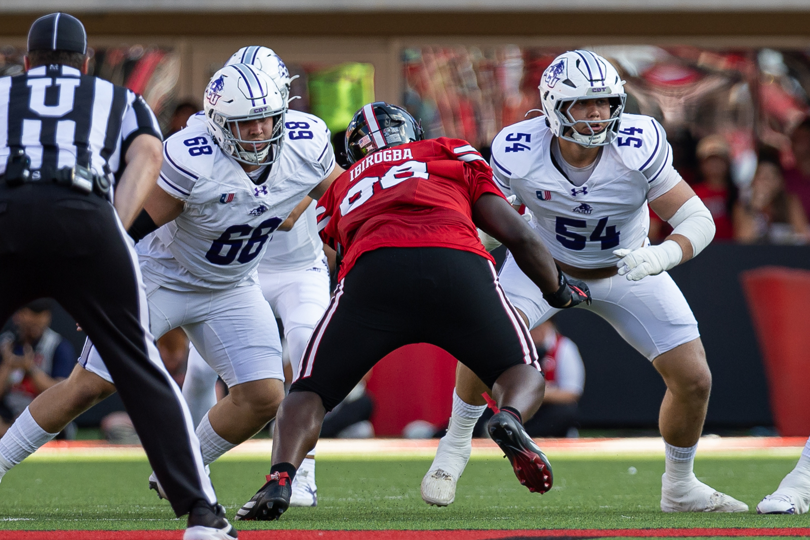 Tech-Abilene-FB-1 | Press Pass Sports Red-jersey running back Ibirogba is sandwiched between two white-jersey defenders as a tackle unfolds on the football field, with a referee nearby.