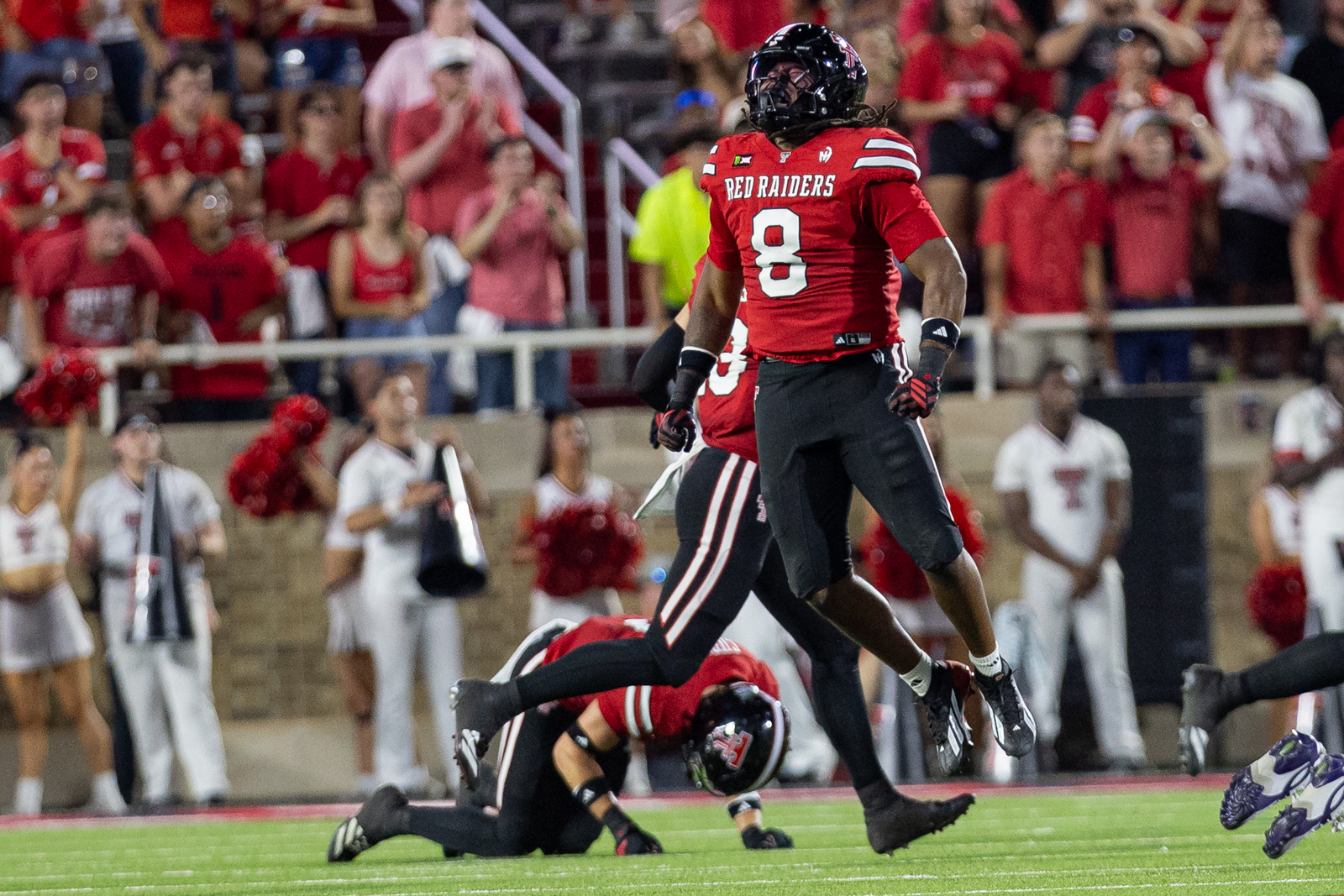 Tech-Abilene-FB-23 | Press Pass Sports Red Raiders player #8 jumps over a fallen opponent on the field during a live game, crowd cheering in the stands behind.