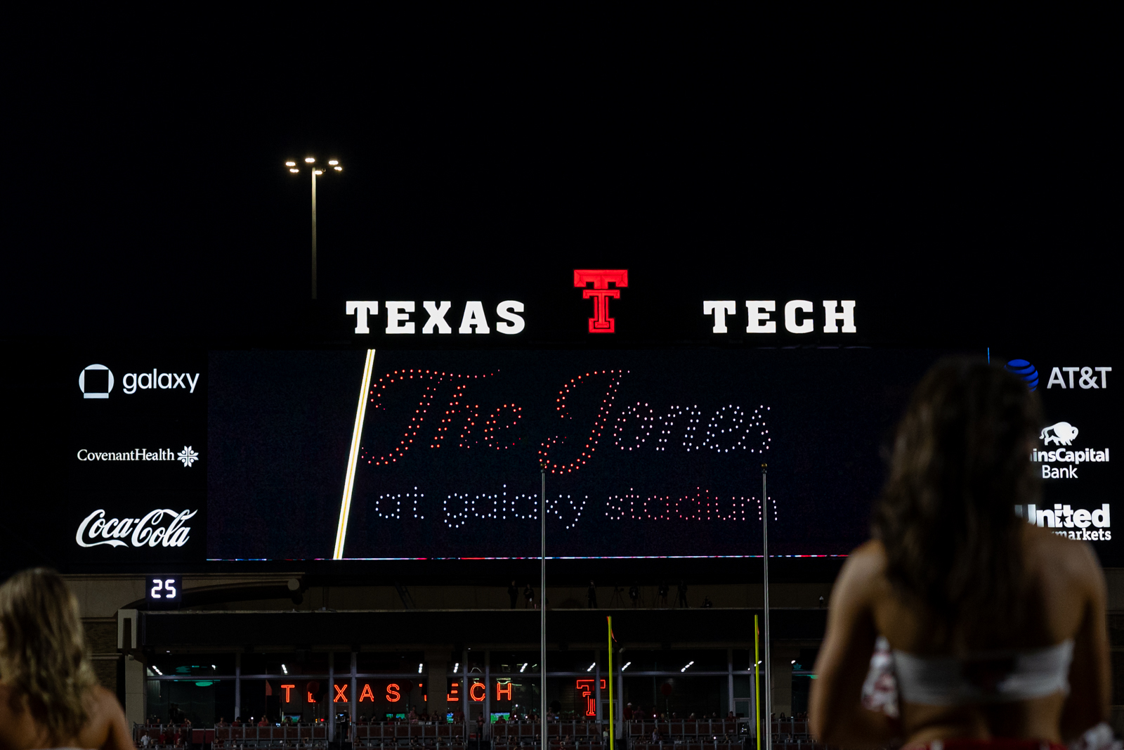 Tech-Abilene-FB-27 | Press Pass Sports Nighttime view of Texas Tech stadium with a bright'TEXAS TECH' sign and sponsor logos along the sides, silhouetted fans in the foreground.