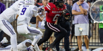 American football player in red jersey (No. 6) runs with the ball as white-clad defenders close in on the field goal line.