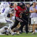 American football player in red jersey (No. 6) runs with the ball as white-clad defenders close in on the field goal line.