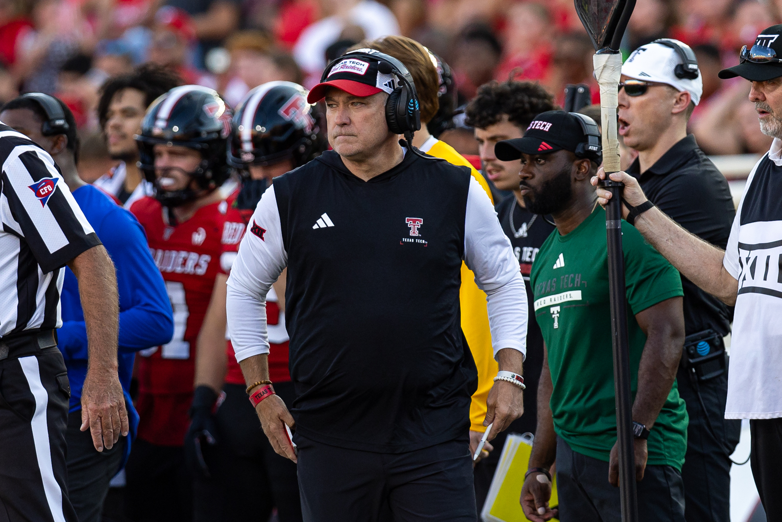 Tech-Abilene-Joey-3 | Press Pass Sports Football coach wearing a headset and Texas Tech outfit stands on the sideline during a game, focused and surrounded by players and staff.