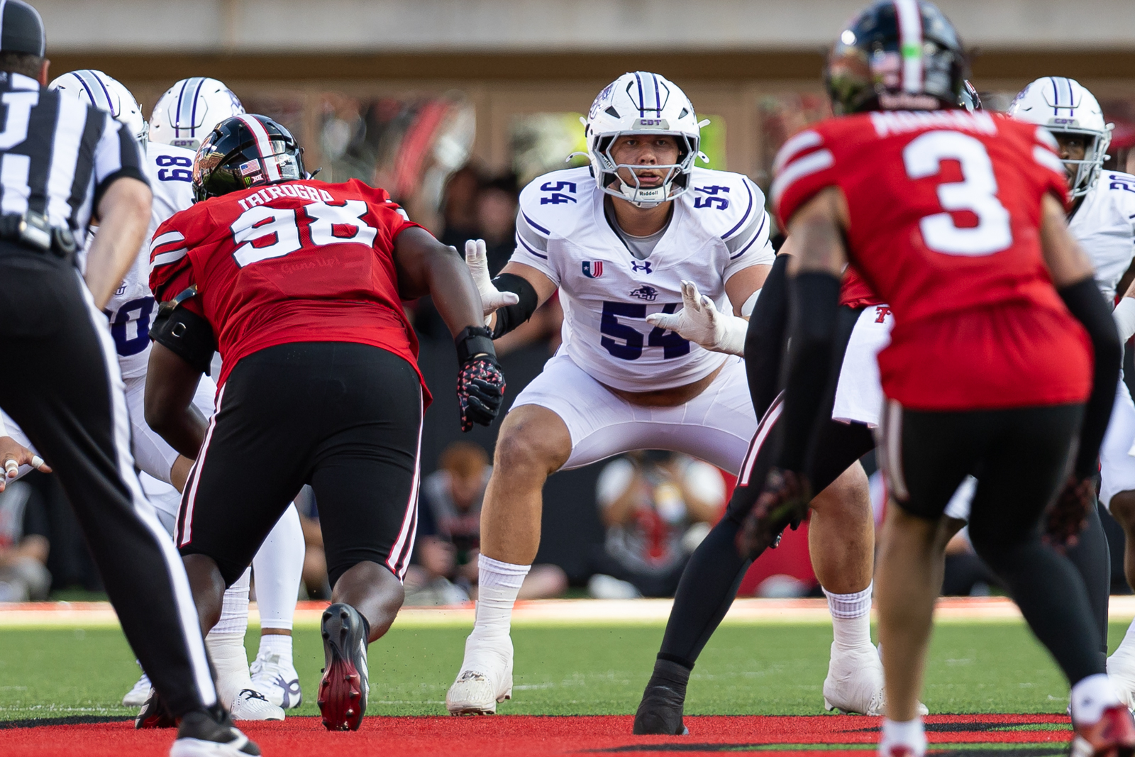 Tech-Abilene-FB-2 | Press Pass Sports White-team lineman wearing #55 crouches at the line of scrimmage facing red-team defenders during a play on the field.