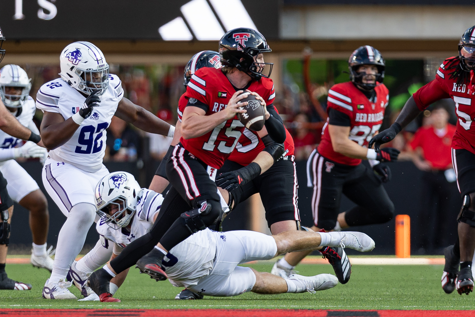 Tech-Abilene-FB-3 | Press Pass Sports Red team quarterback wearing #15 cradles the football while tackling blocks from white-uniformed defenders during a college football game on a green field.