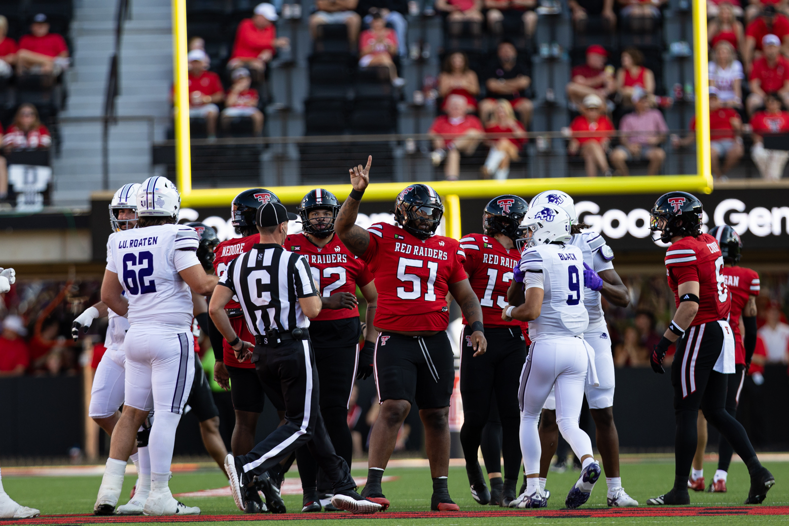 Tech-Abilene-FB-4 | Press Pass Sports Group of Red Raiders players gather on the football field, with a referee nearby and a yellow goalpost in the background during a game.