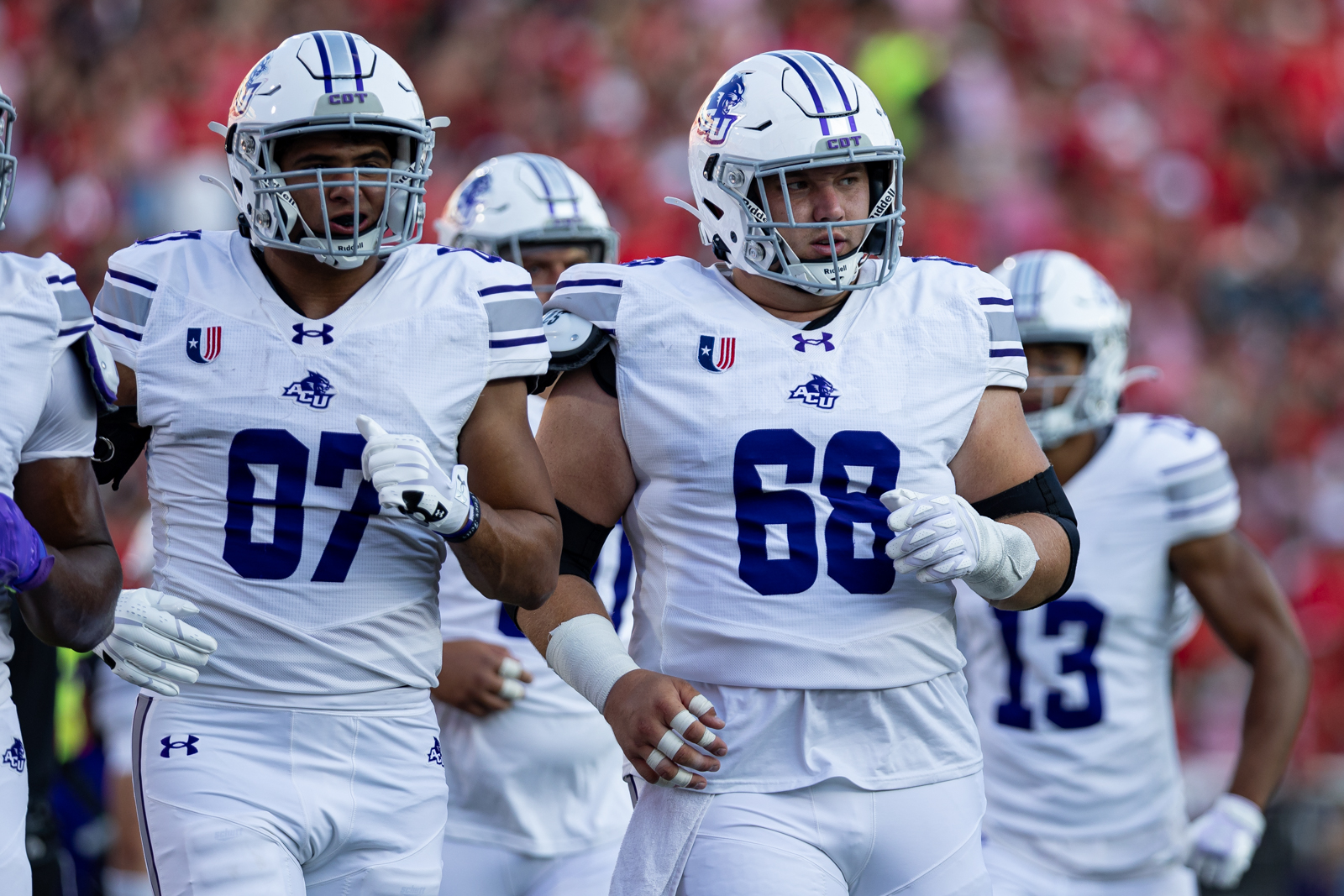 Tech-Abilene-FB-7 | Press Pass Sports Two football players in white uniforms with blue numbers 07 and 68 run together on the field.