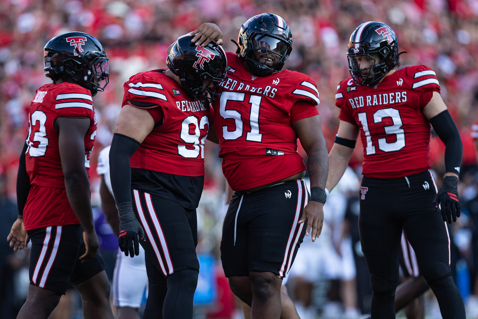 Tech-Abilene-FB-8 | Press Pass Sports Four Texas Tech football players in red uniforms celebrate on the field, arms around each other mid-huddle.