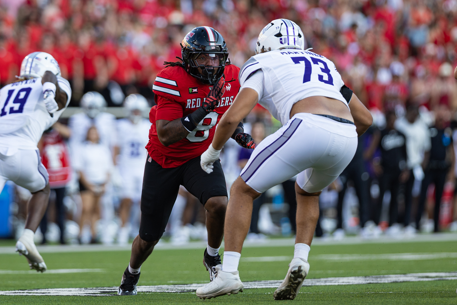 Tech-Abilene-FB-9 | Press Pass Sports Football player in a red Red Raiders jersey rushes toward a defender in white on a green turf with a packed crowd behind them.