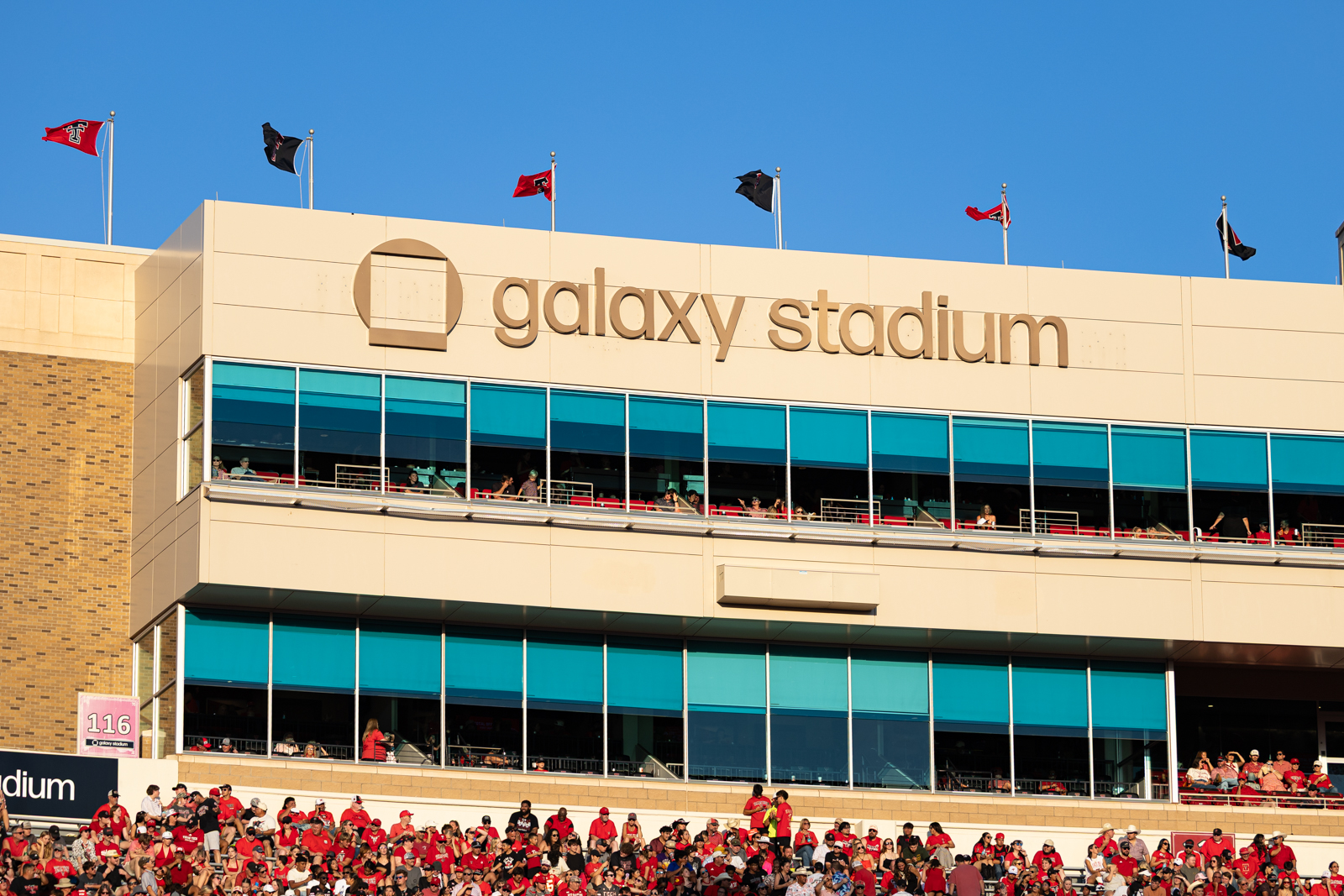 Tech-Abilene-FB-12 | Press Pass Sports Galaxy Stadium exterior with tan facade, blue-tinted windows, and flags on the roof under a clear blue sky, with a red-clad crowd in the stands below.