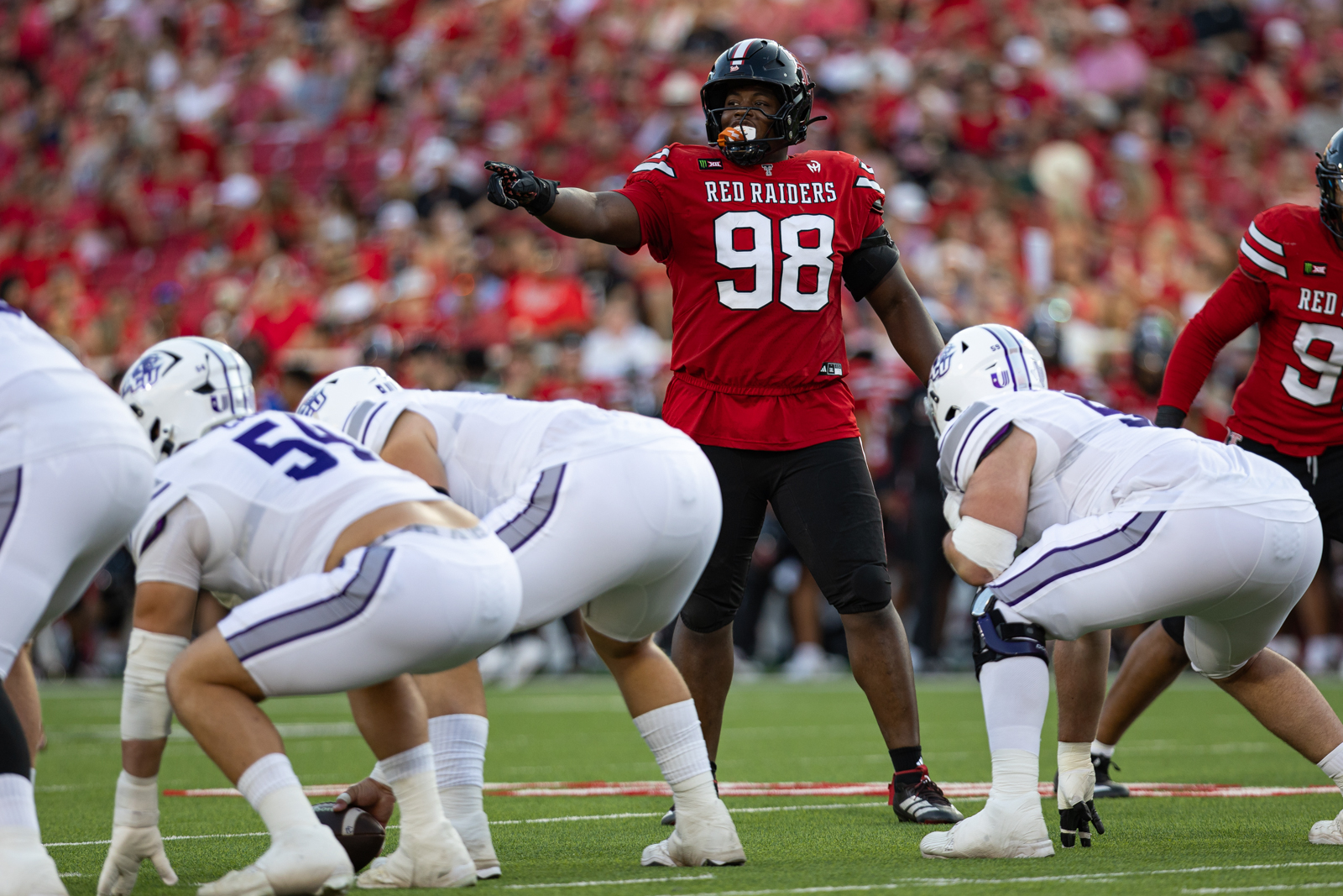 Tech-Abilene-FB-14 | Press Pass Sports Red Raiders lineman wearing #98 points toward the offense at the line of scrimmage during a football play with white-uniformed defenders nearby in a packed stadium.
