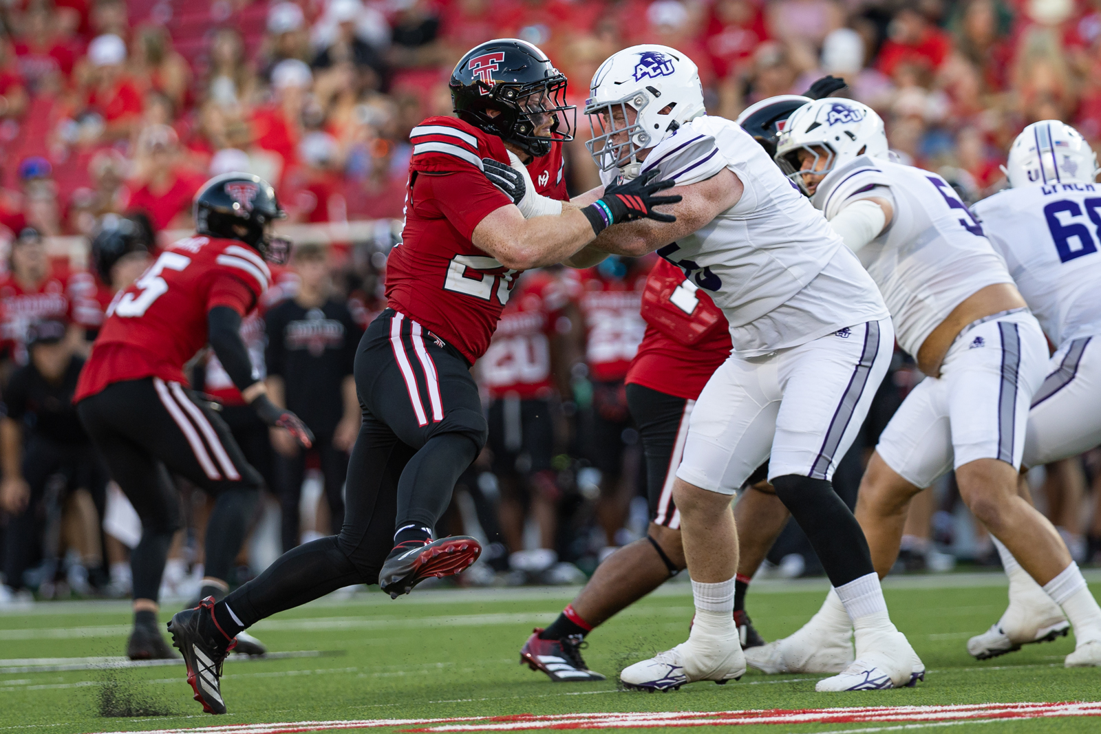 Tech-Abilene-FB-15 | Press Pass Sports Red-clad defender and white-clad opponent collide at the line of scrimmage on a green football field as the crowd watches in the background.