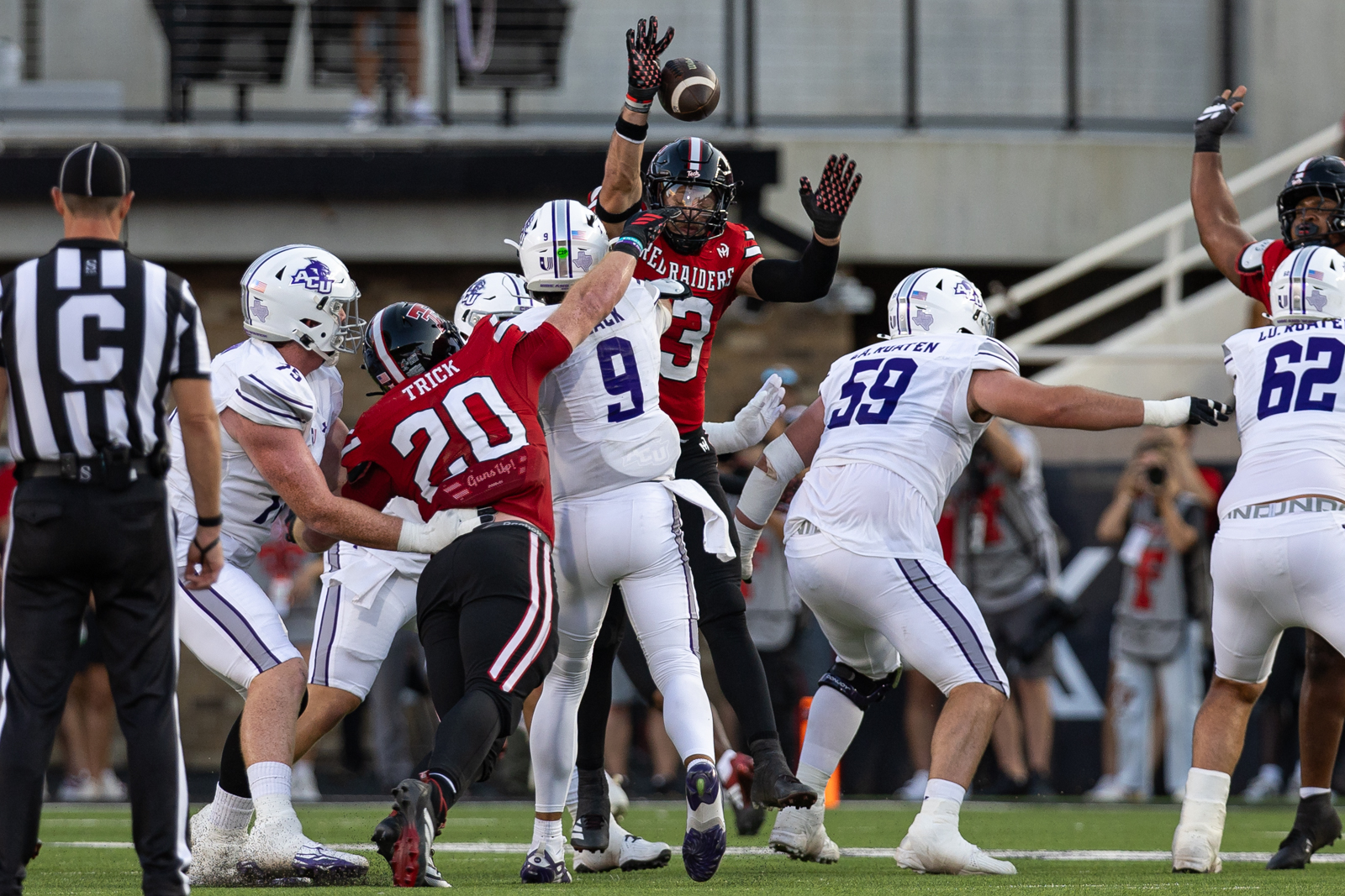 Tech-Abilene-FB-16 | Press Pass Sports Players leap for a football at the line of scrimmage; a white-uniformed defender reaches for the ball as a red-clad opponent blocks nearby.