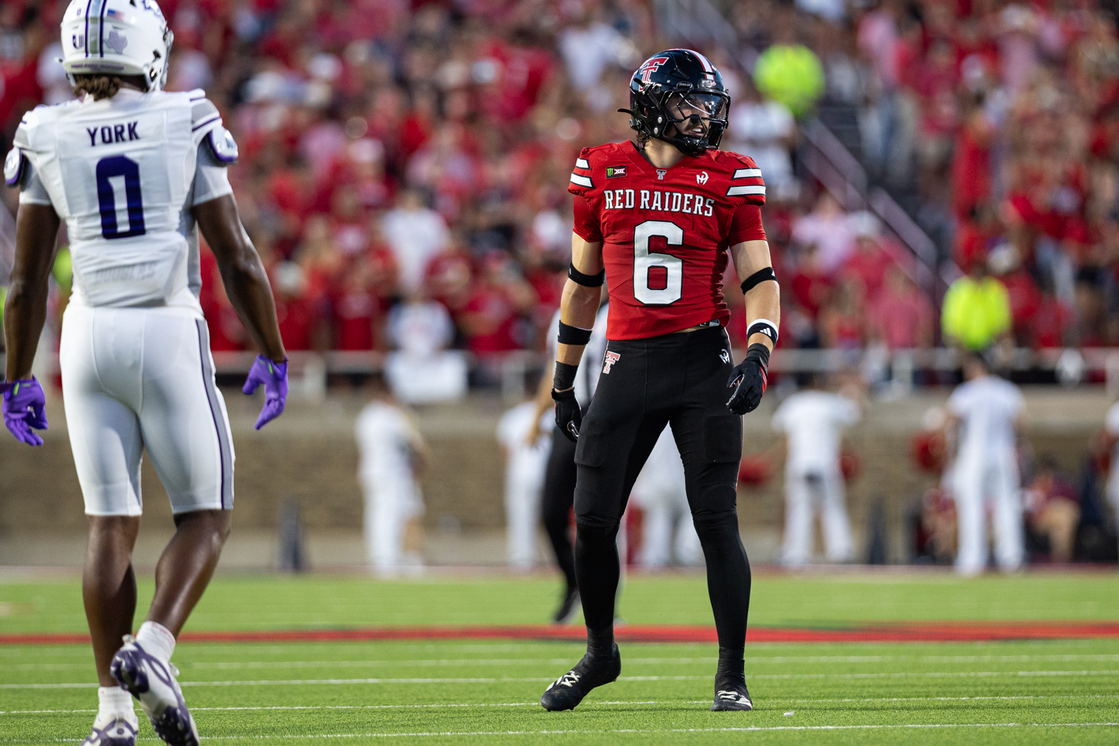 Tech-Abilene-FB-20 | Press Pass Sports Football player in a red Texas Tech Red Raiders jersey (#6) stands on the field as a white-clad opponent approaches; crowd in the stadium in the background.