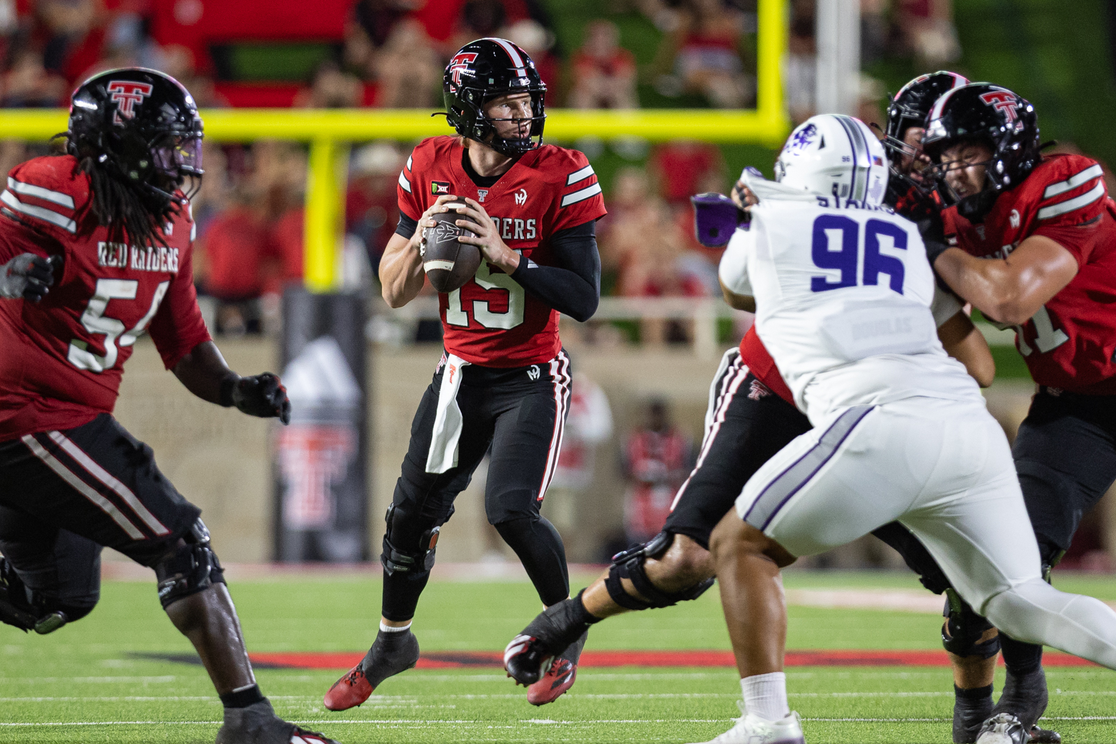 Tech-Abilene-FB-25 | Press Pass Sports Quarterback in a red Red Raiders jersey (#15) prepares to pass as defenders close in on a night game field.