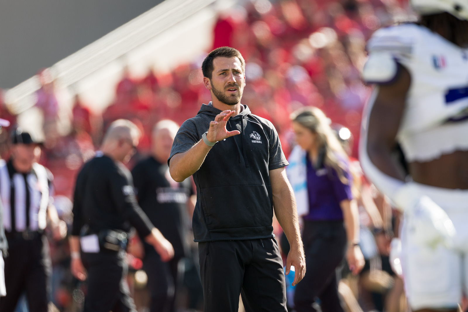 abilene-christian-vs-texas-tech-1 | Press Pass Sports Coach in a dark hoodie signaling with his hand on a football field, crowd in red stands behind him