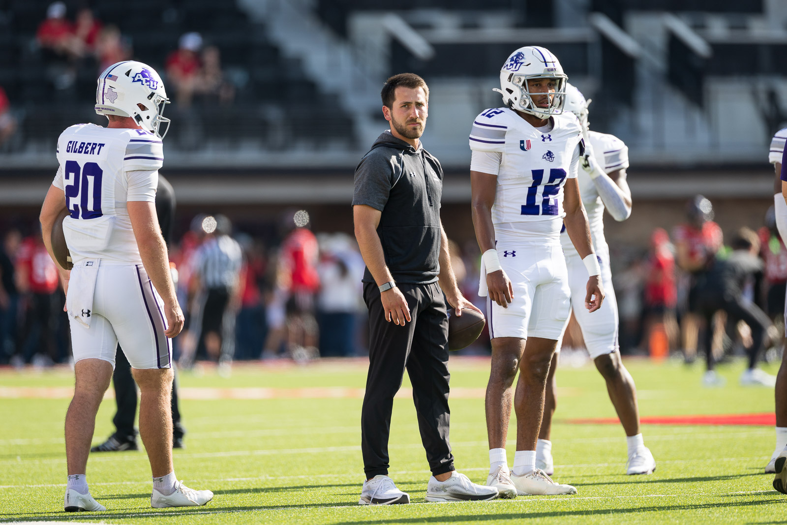 abilene-christian-vs-texas-tech-3 | Press Pass Sports Coach in dark gray hoodie stands with two football players in white uniforms on a sunny practice field.