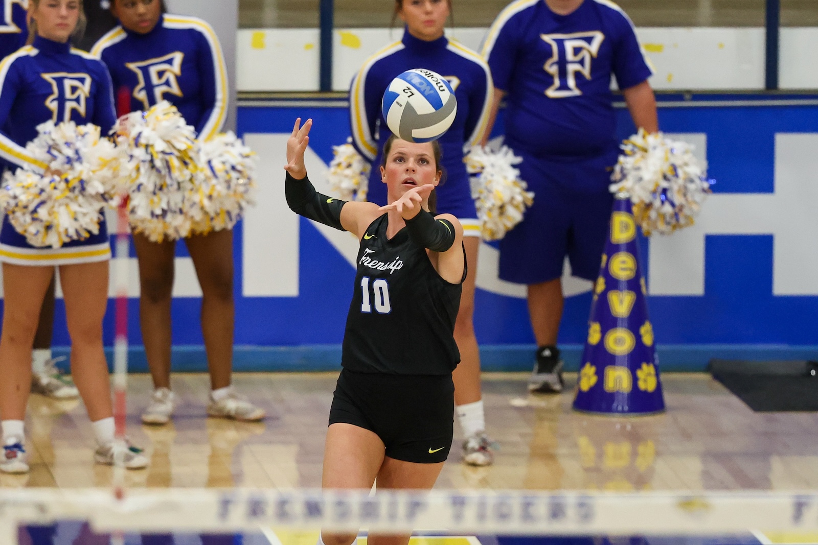 Seabolt Chase-11790 | Press Pass Sports Female volleyball player wearing a black #10 uniform prepares to pass the ball, with cheerleaders in purple uniforms watching in the background.