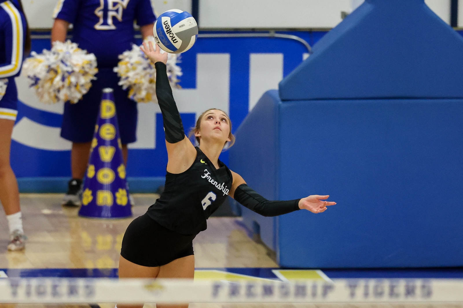 Seabolt Chase-11937 | Press Pass Sports Female volleyball player in a black uniform (number 5) serves or sets the ball during a match in a gym with blue padding and cheerleaders in the background