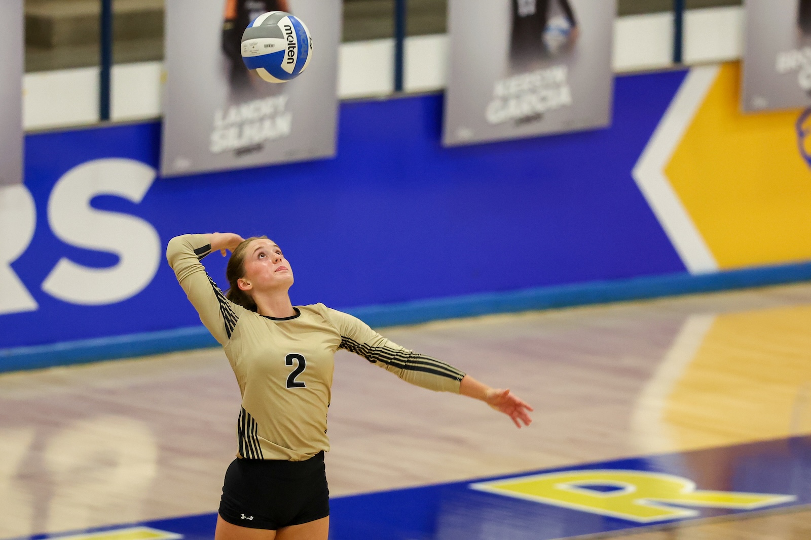 Seabolt Chase-12095 | Press Pass Sports Female volleyball player in beige jersey number 2 serves the ball on an indoor court.
