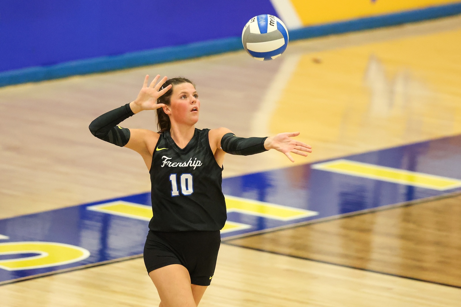 Seabolt Chase-12388 | Press Pass Sports Female volleyball player in a black jersey with number 10 reaches to set a ball mid-air in a gymnasium.