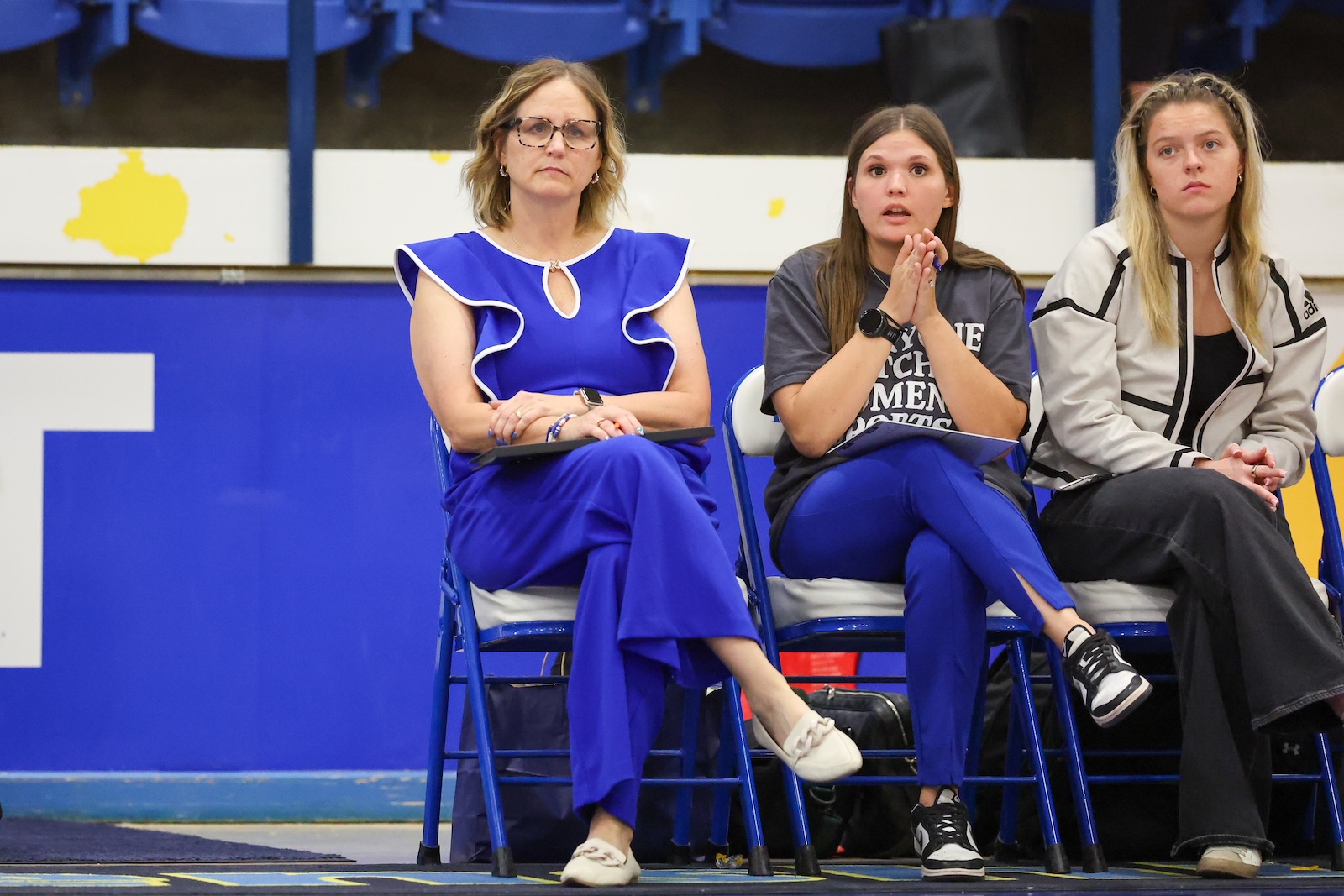 Seabolt Chase-12643 | Press Pass Sports Three women sit on blue folding chairs in a gym, from left: a woman in a bright blue dress, a young woman in a gray T‑shirt, and a woman in a white jacket.